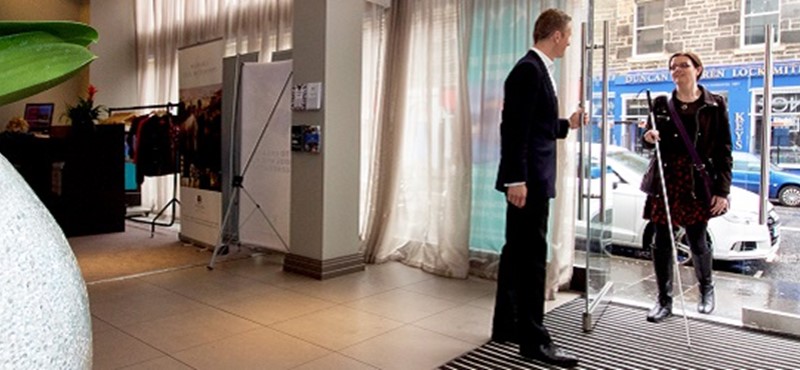 Photo of a hotel staff member meeting a long cane user at the front entrance.