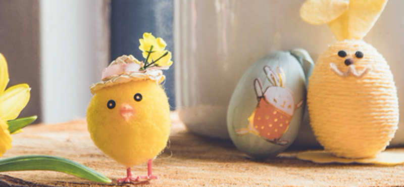 image of an egg painted to look like a bunny sitting next to a small plastic chick and yellow flower on a wooden table. The table is against a bright window.