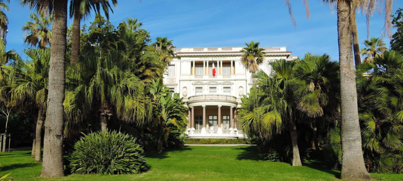 Exterior image of Musée Masséna, there are many palm trees in the garden in front of the building.