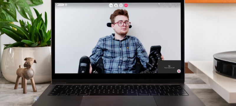 Photo of Jamie Hale appearing on a video call on a laptop. The laptop is on a table next to a house plant and a wooden dog ornament.