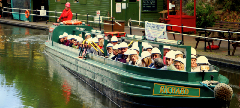 A green narrowboat filled with people wearing white helmets on a canal.