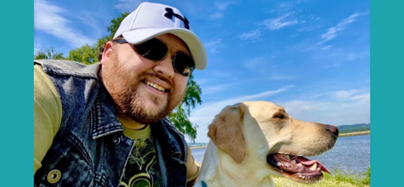 A picture of Jonathan wearing sunglasses and a cap next to his guide dog Sam. A blue sky can be seen in the background of the image. The image is against a teal background. 