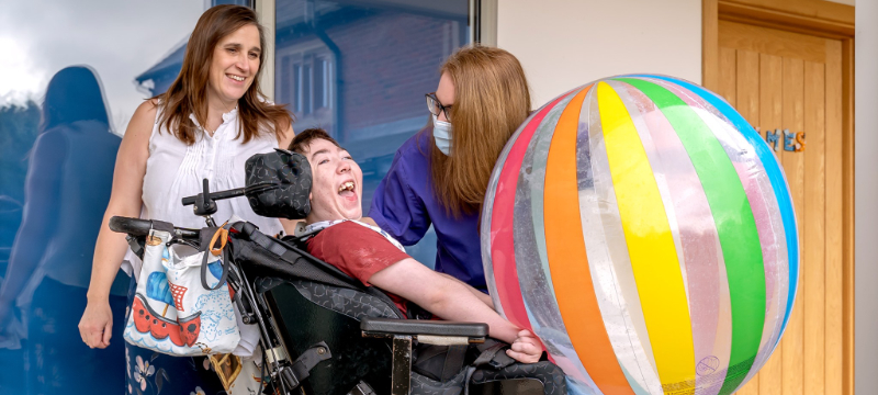 James smiling in the centre of the image with a large colourful ball. Christine (left) and Helen (right) pictured behind him.