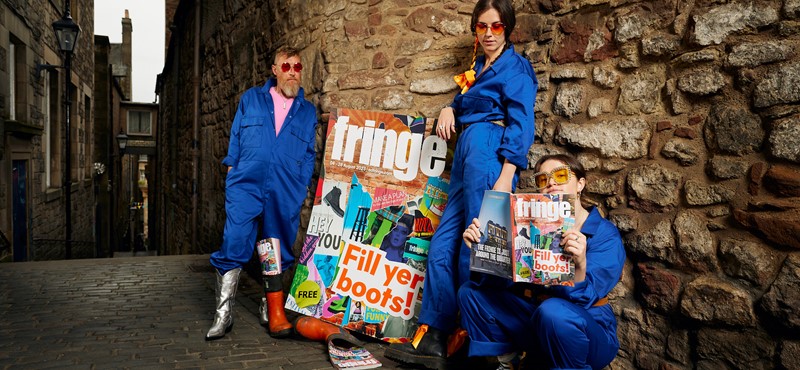 Three people wear cobalt blue boiler suits while posing in a lane holding Edinburgh Festival Fringe promotional poster