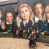 Photo of me sitting in a powerchair infront of a mural of the Derry Girls in Derry, Northern Ireland.