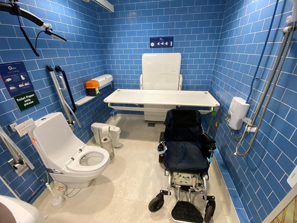 A closer view of the adult changing and washing table, with a powerchair parked in front of it, with ample room. There is a shower, a hoist, a shelf with sharps box, toilet roll holder, sanitary bin, and instructions.