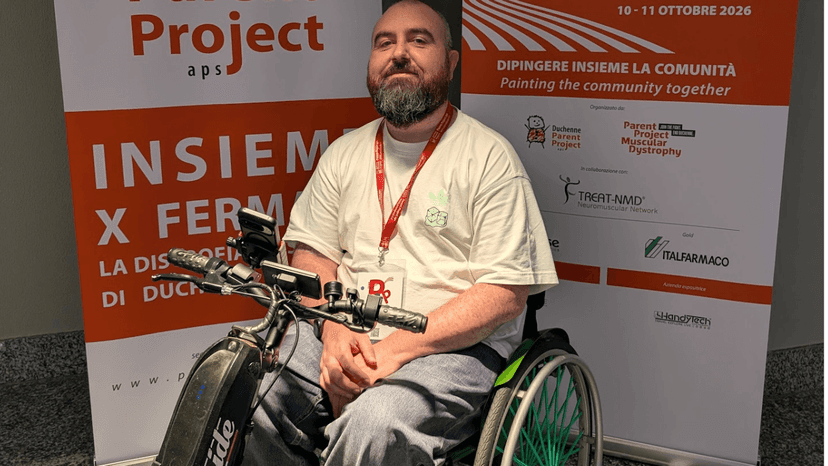 Joe a wheelchair user, sits in front of two banners promoting the Becker Muscular Dystrophy event in Milan