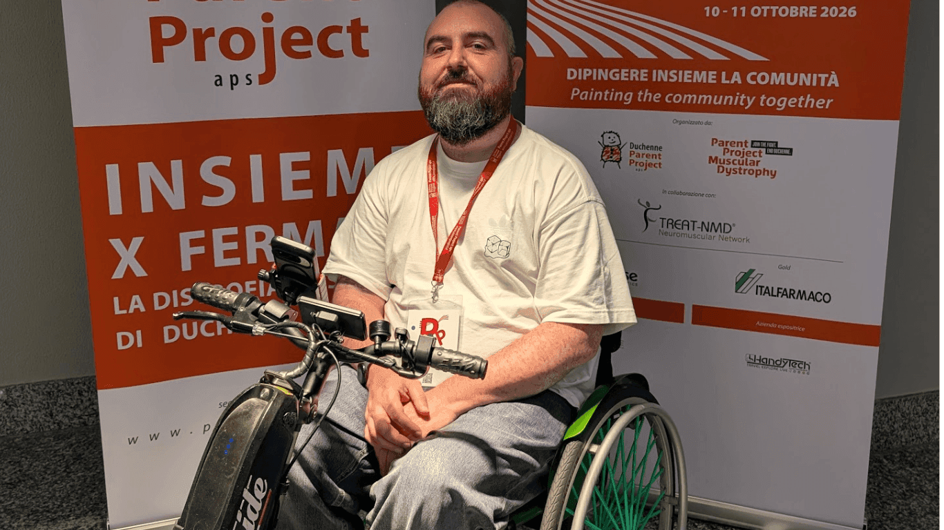 Joe a wheelchair user, sits in front of two banners promoting the Becker Muscular Dystrophy event in Milan