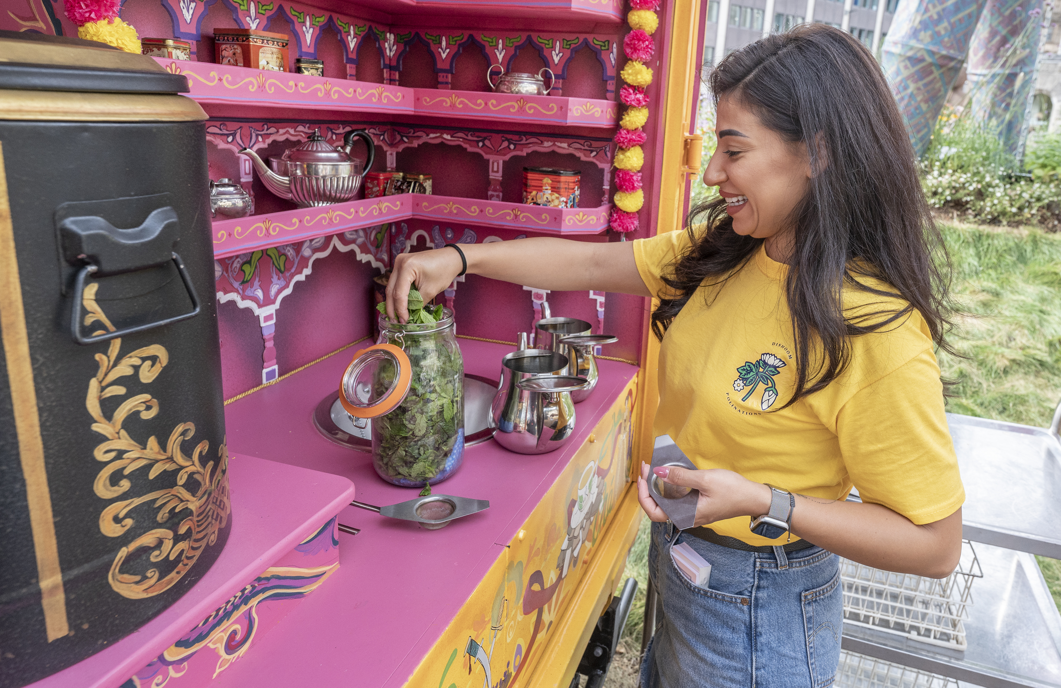A woman in a yellow t shirt and jeans stands at a pink stall as she takes mint tea leaves out of a glass jar