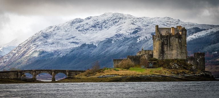 Photo of Eilean Donan Castle with mountains on the background and surrounded by a lake.