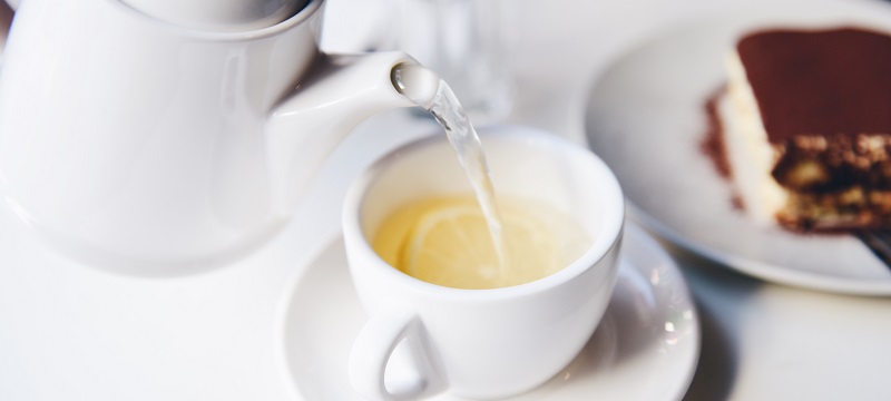 Photo of tea pot pouring hot water in a tea cup with slices of lemon.