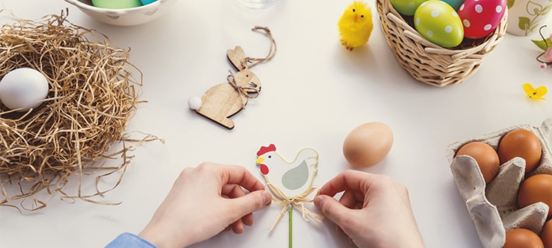 Image of hands creating an Easter craft - a cut out of a cartoon chicken on a stick. Also on the table is a nest, some eggs and a wooden bunny rabbit.