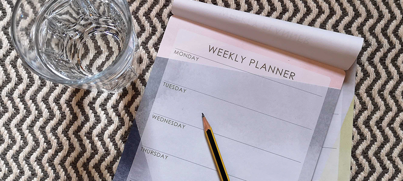 Image of a weekly planner with a pencil on top and a glass of water beside it and a patterned background