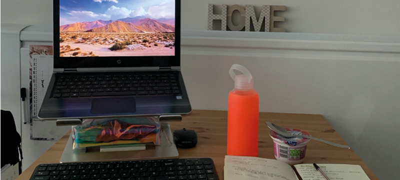 Image of a laptop on a raised stand with a keyboard, pencil case and computer mouse underneath. Also on the table is a book, water bottle and tub of yogurt.