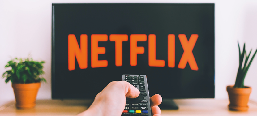 Image of a hand pointing a TV remote at a TV screen that has the Netflix logo in red.