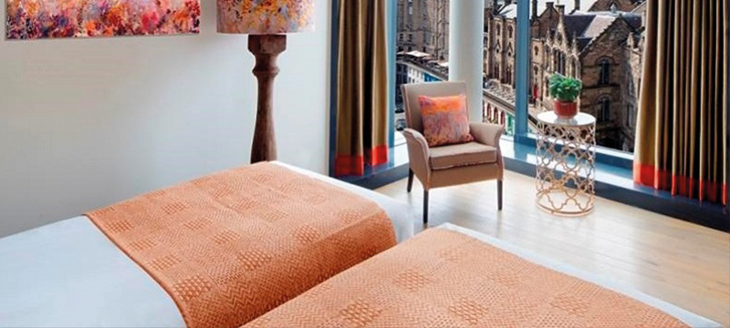 Two beds next to each other in a hotel bedroom with a corner window and space at the end of the beds.