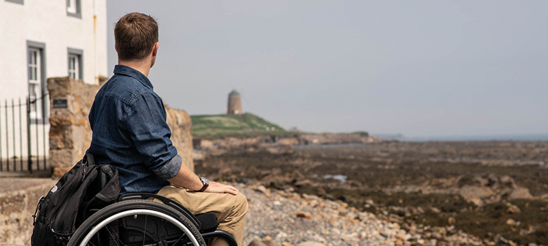 A man looks out over a rocky beach. He is wearing a blue shirt, pale yellow trousers and sitting in a manual wheelchair.