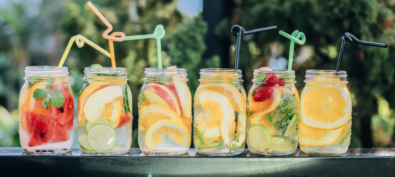 A line of drinks in glass jars with fruit garnishes and plastic straws.