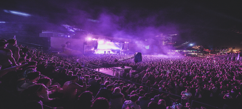 Image of a concert, with an overview of the whole crowd tinged purple with lights.