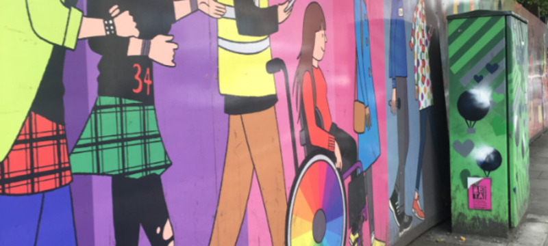 Artwork on a wall of St. Stephen’s Green in Dublin, Ireland showing a young wheelchair user dressed in red and black rolling alongside different people of varying identities. The spokes guard of the wheelchair has rainbow colours.