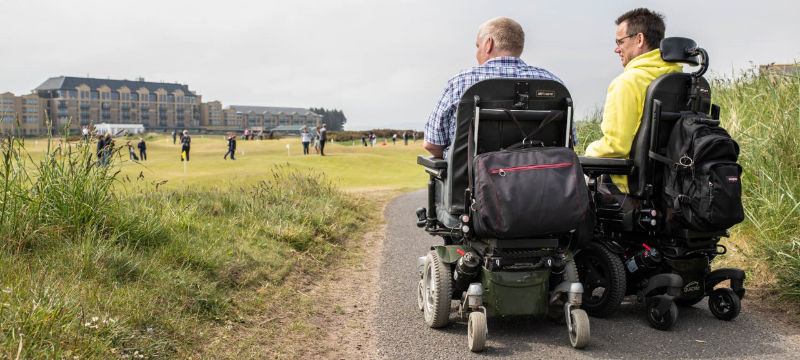 A golf course with two powerchair users pictured with their backs to the camera on a path at the right side of the image.