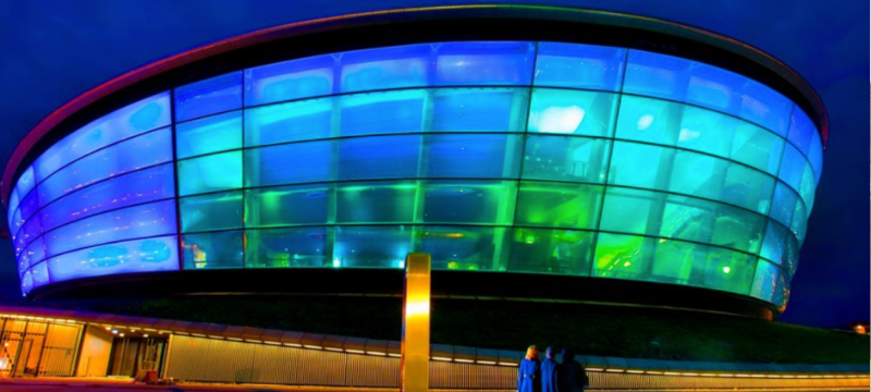 A large circular building lit up with blue and green lights. Three people stand looking up at the building.