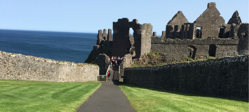 A path leading down to an ancient castle with the sea in the background. There is grass either side of the path.