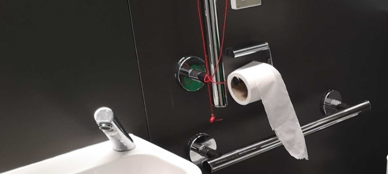 Inside of an accessible toilet with black walls, silver grab rails and a white sink. The toilet roll is in a holder attached to the wall. The red emergency cord has been wrapped around one of the grab rails making the cord and grab rail hard to use.