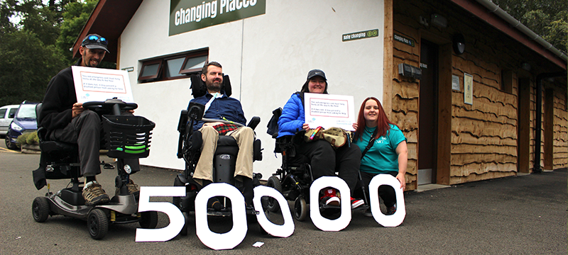 Four people pictured outside a Changing Places toilet. They are holding giant Red Cord Cards and the number 50,000 can be seen at their feet.