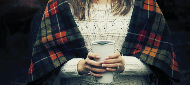 A woman with a tartan blanket around her shoulders. She is wearing a light jumper and holding a flask for a hot drink.
