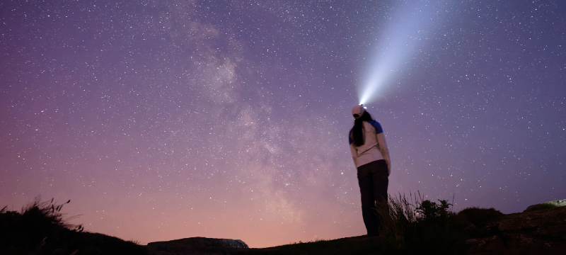 A woman with her back to the camera. She has a head torch on and is looking up at a purple sky filled with stars.