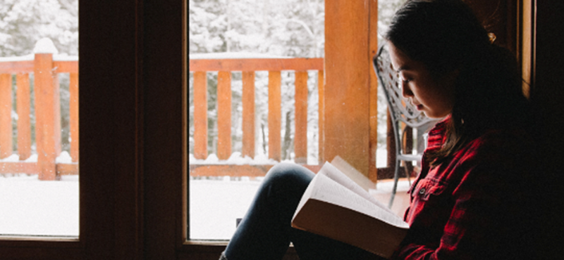 A woman reading a book inside. She is on the ground beside a glass door.