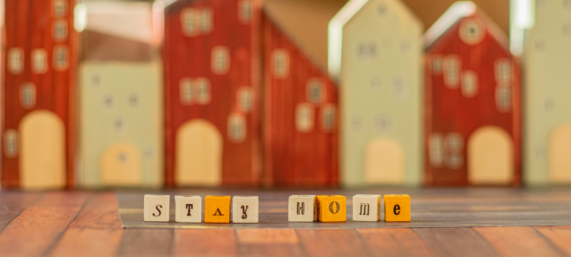 "Stay home" written in cube letters. Blurred outlines of miniature buildings can be seen in the background.