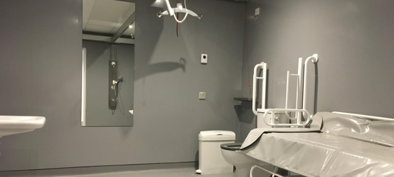 The Changing Places has grey walls. A sink can just be seen on the left of the image. There is a large mirror on the back wall with a hoist seem overhead. On the right we can see a bin, toilet and adult sized changing bench.