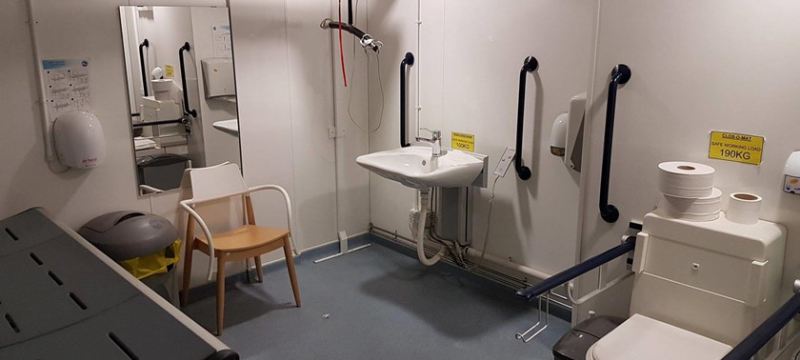An adult sized changing bench on the left with a bin, chair and mirror next to it. A hoist can be seen in the corner of the room next to the sink. The toilet is pictured in the right corner.