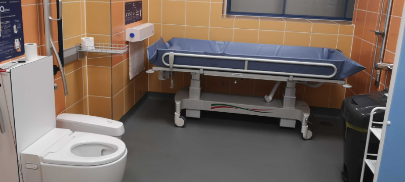 Inside a Changing Places toilet with orange tiles on the walls. The picture includes a toilet on the left and wide tear off paper roll and an adult sized changing bench in the centre.