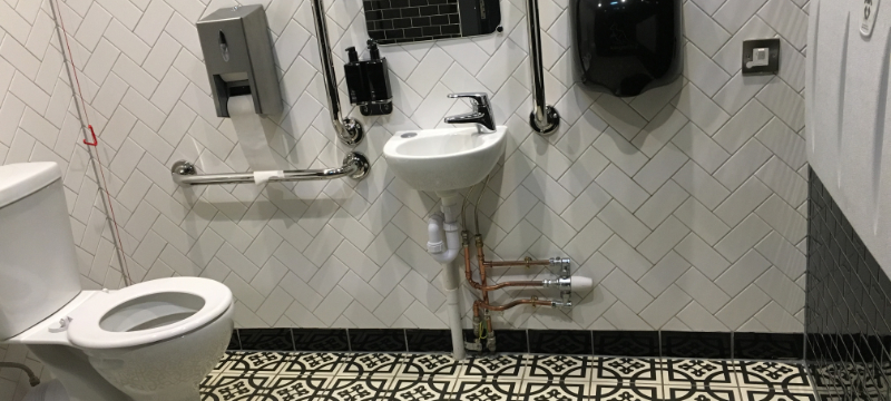 Toilet pictured on the left with a patterned tile floor and white tiles on the wall. There is a small sink in the centre of the image with a hand dryer pictured next to it. On the right is a baby changing table and black tiles below it.