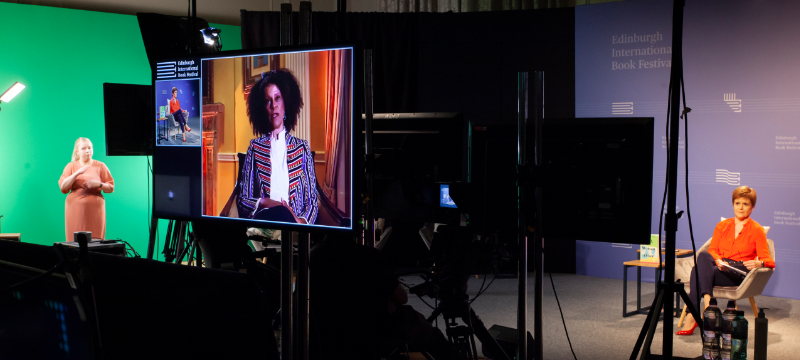 A BSL interpreter is pictured in front of a green screen at the left of the image. A large screen is beside her showing two people on a video call together, one of the people on the screen is also pictured on the right of the image.