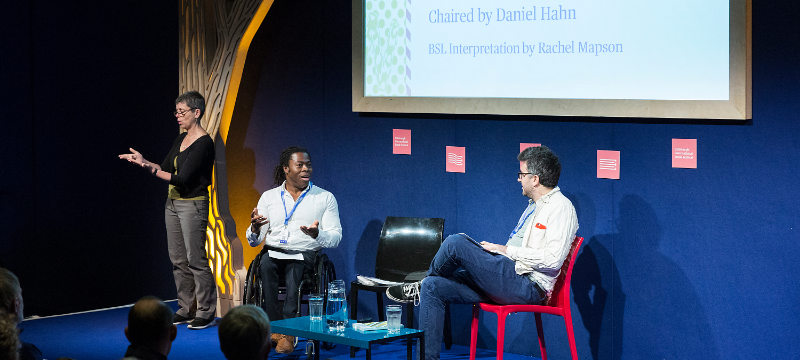 Three people on stage at an event. One on the left is standing, one in the centre is seated in a wheelchair and one on the right is sitting on a chair. A large screen can be seen behind them.