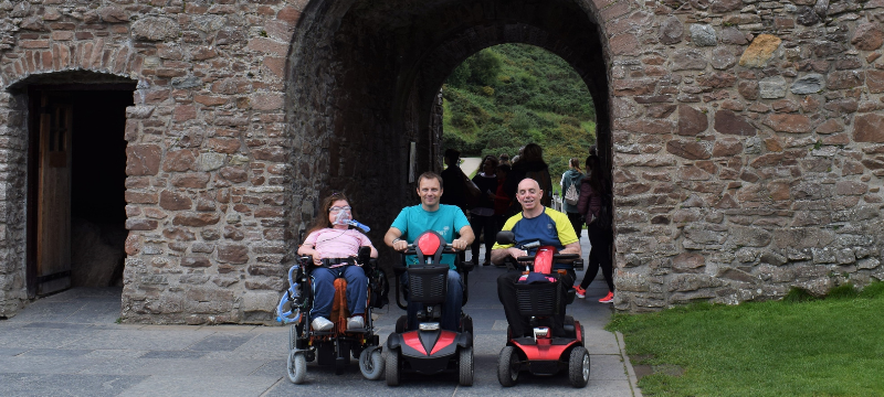 Karis, Ryan and Gordon pictured next to each other in front of an old brick building. Karis is in a powerchair. Ryan and Gordon are using scooters.