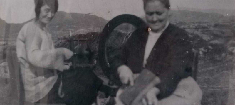 Old black and white photo with two woman pictured sitting on seats outside spinning wool.