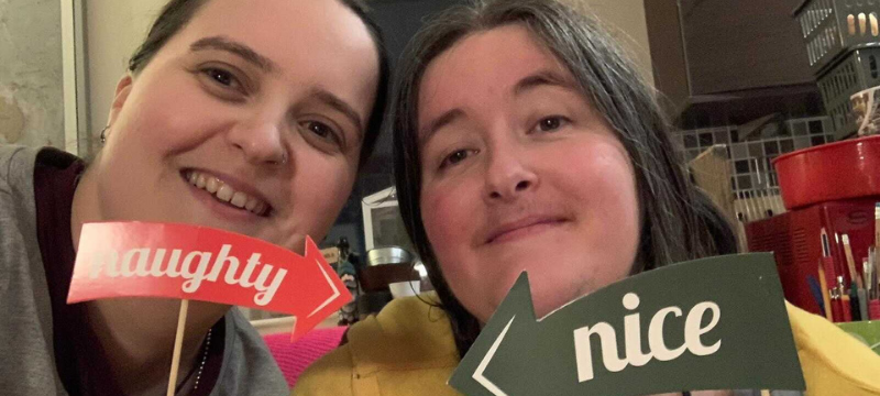 Sarah on the left with a 'naughty' sign pointing to Karine. Karine is on the right with a 'nice' sign pointing to Sarah.