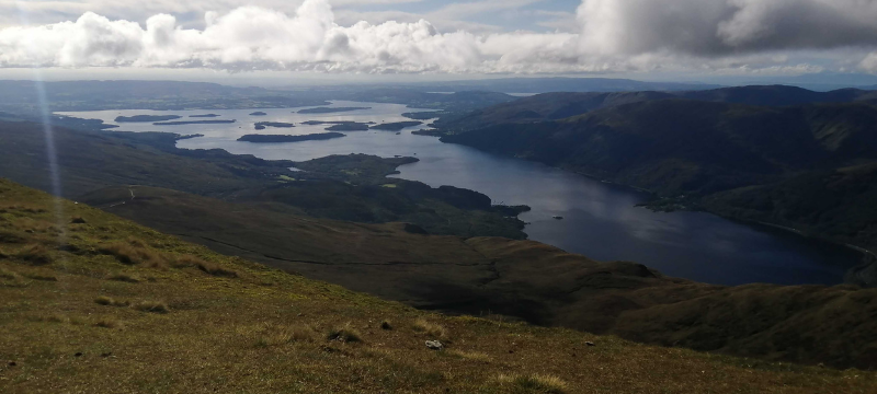 Image taken from a hill looking down at a loch with clouds seen in the sky above.