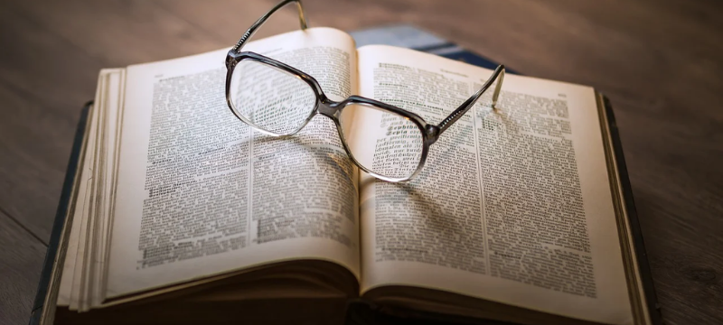 Image of a book sitting on a wooden surface and a pair of glasses sitting on top of the book.