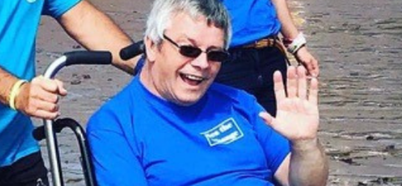 Image of Neil sitting in a beach wheelchair waving to the camera.