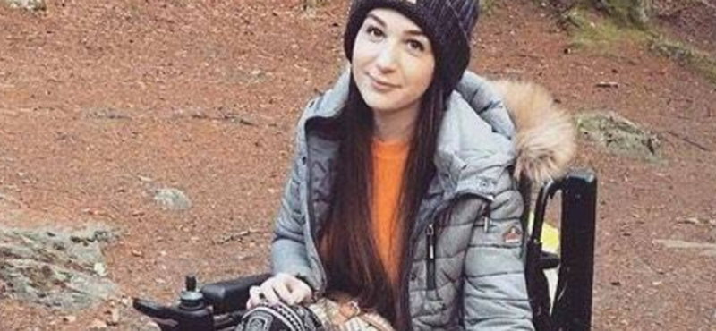 Image of Hollie sitting in her wheelchair smiling at the camera with open woods behind her.