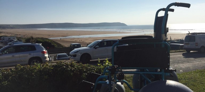 Image of a beach wheelchair sitting on a grass embankment in front of a road with cars and the beach behind them.