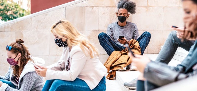 A photograph of students wearing face masks as they sit on stone steps looking at their mobile phones.