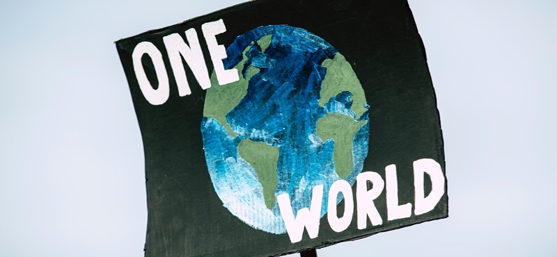 Text reads 'One World' on a black placard which someone is holding up, featuring a globe underneath the text.
