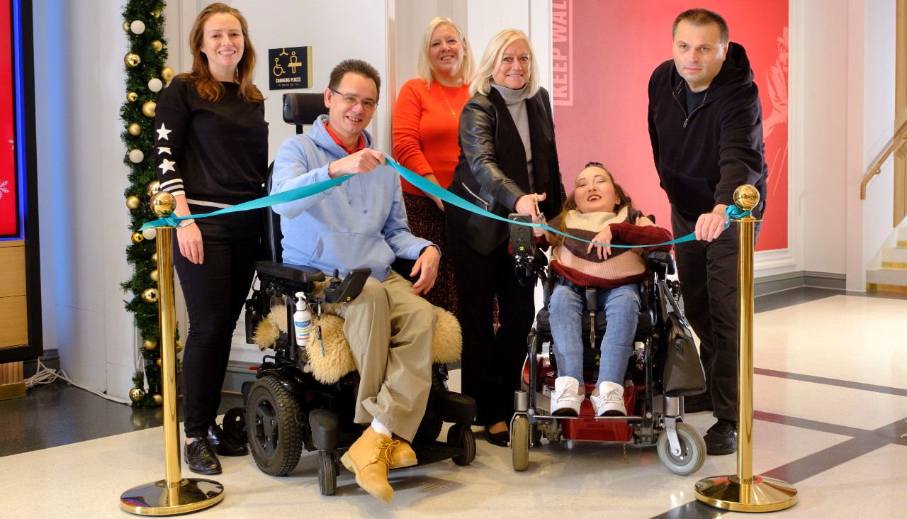 Six people outside the Changing Places Toilet at Johnnie Walker Princes Street with two holding the cyan ribbon at either side and two holding a pair of scissors ready to cut in the middle.